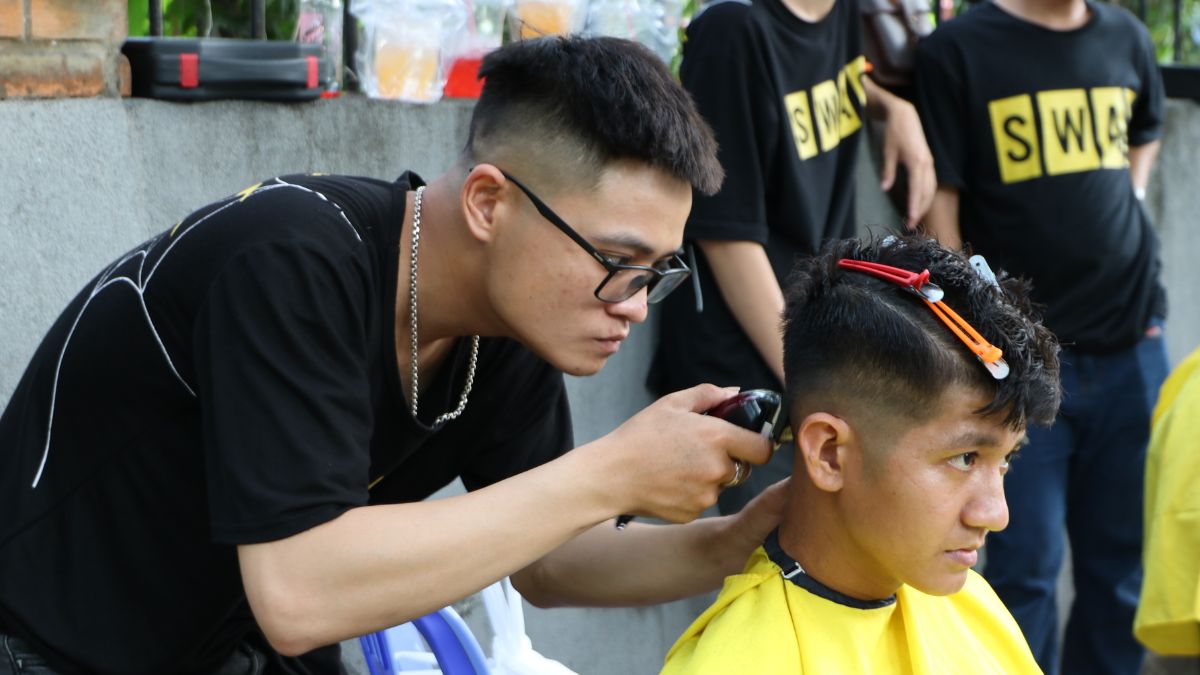 Người thợ cắt tóc đang tập trung dùng tông đơ để cắt tóc cho khách hàng ngồi trên ghế, xung quanh có vài người đứng.
