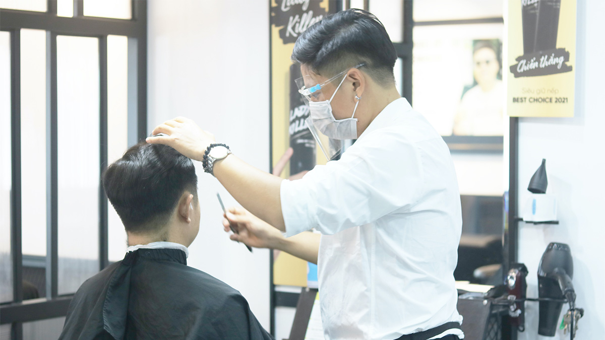 Người thợ cắt tóc đang tỉa tóc cho khách hàng trong tiệm, cả hai đều đeo khẩu trang.