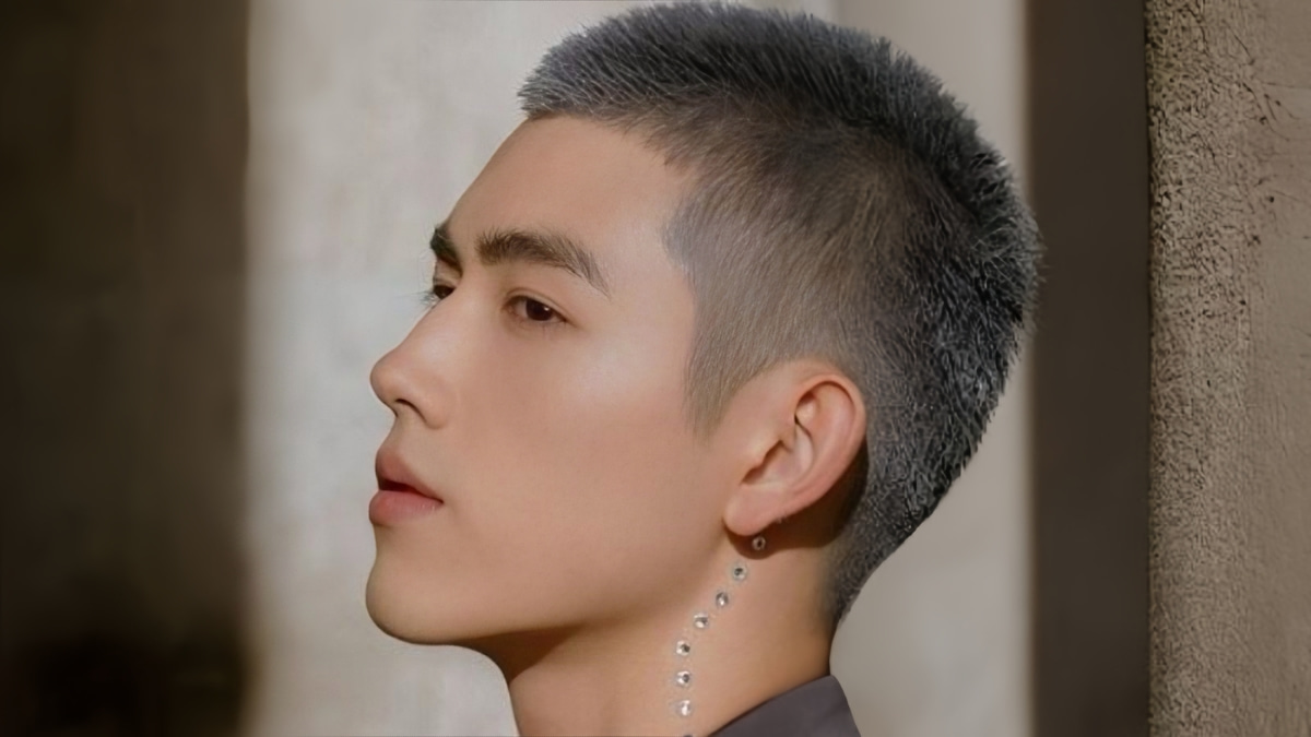 Người đàn ông nhìn nghiêng, tóc ngắn, có khuyên tai và các phụ kiện nhỏ trên cổ, nền mờ.