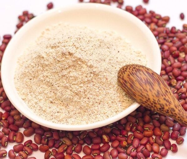 Bột trong bát trắng, bao quanh bởi hạt đậu đỏ và muỗng gỗ nâu, trên nền trắng.