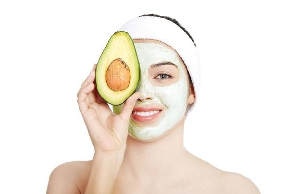 Người với mặt nạ dưỡng da, khăn quấn tóc, cầm nửa quả bơ đặt trước mắt, nền trắng.