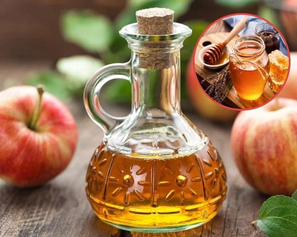 Chai thủy tinh đựng chất lỏng màu vàng nâu, xung quanh có táo tươi, ảnh nhỏ bên trong có mật ong.