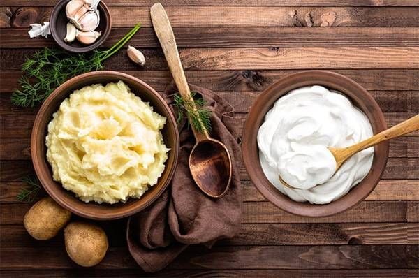 Bát khoai tây nghiền và bát kem chua trên bàn gỗ, kèm thìa gỗ và rau thơm, tỏi, khoai tây trang trí xung quanh.