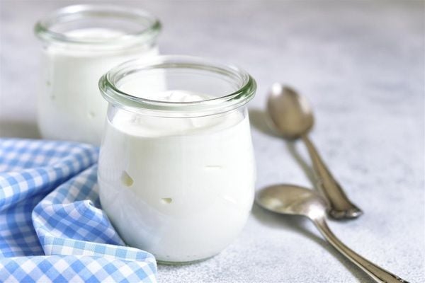 Hai hũ sữa chua thủy tinh trên bàn, bên cạnh là hai chiếc thìa và khăn kẻ caro xanh trắng.