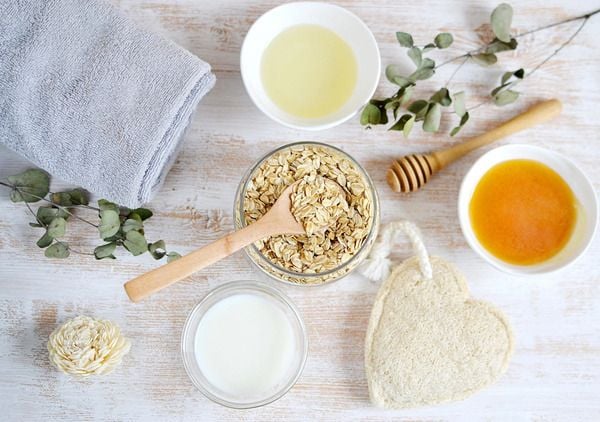 Khăn tắm, bột yến mạch, mật ong, dầu, và đồ trang trí trên nền gỗ sáng.