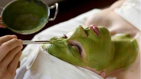 Người đang được đắp mặt nạ màu xanh lá cây lên mặt bằng cọ, nằm thư giãn với khăn trắng quấn quanh đầu.