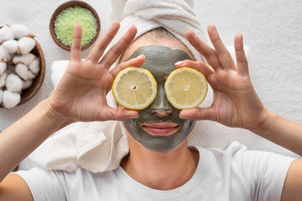 Người đắp mặt nạ bùn thư giãn, tay cầm hai lát chanh che mắt, khăn quấn đầu, nằm trên khăn tắm.