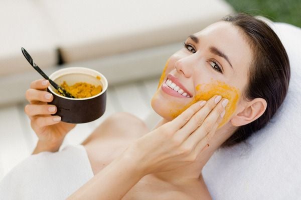 Người phụ nữ nằm thư giãn, thoa mặt nạ nghệ lên mặt, cầm chén nhỏ màu đen.