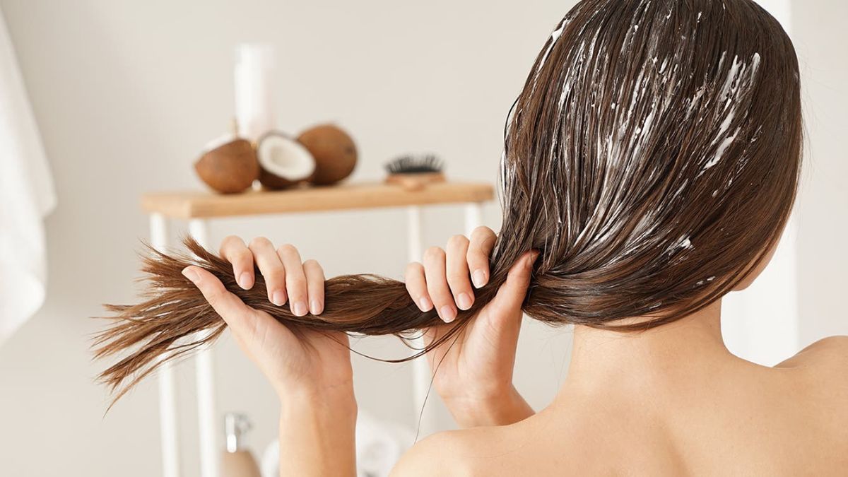 Người đang dưỡng tóc với sản phẩm dưỡng trắng, nền có dừa và bàn chải trên kệ gỗ.