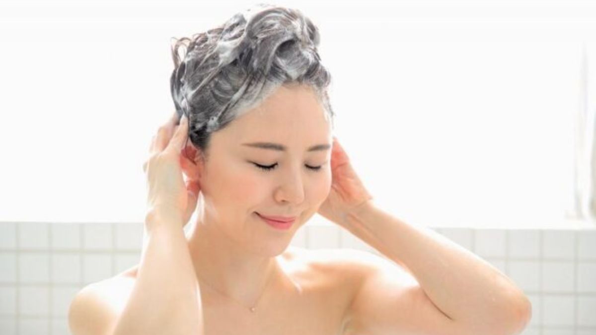 Người đang gội đầu với bọt xà phòng, mắt nhắm và biểu cảm thư giãn, nền gạch trắng.