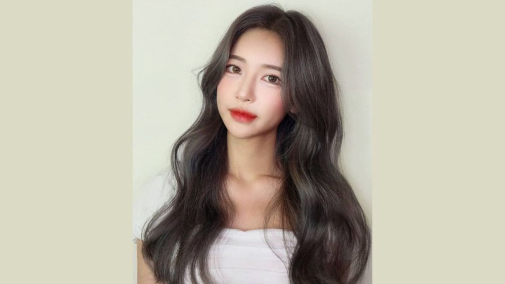 Người phụ nữ với mái tóc dài màu nâu sẫm, mặc áo trắng, nhìn thẳng vào máy ảnh trên nền màu trung tính.