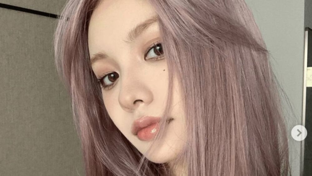 Người với mái tóc dài màu tím pastel đang nhìn về phía máy ảnh trong không gian trong nhà.