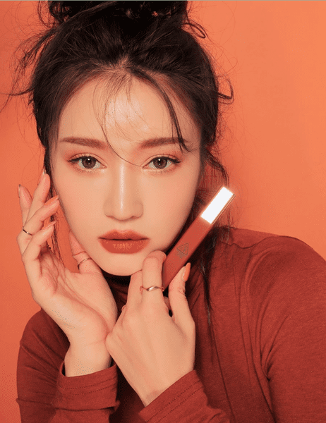 Người cầm sản phẩm trang điểm màu cam, nền cam, ánh sáng tập trung vào gương mặt và sản phẩm.
