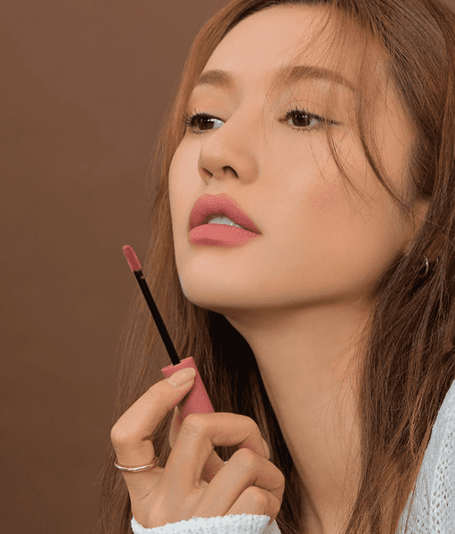 Người phụ nữ cầm cọ son môi màu hồng, nền nâu, ánh sáng dịu nhẹ, tạo dáng nghiêng mặt.