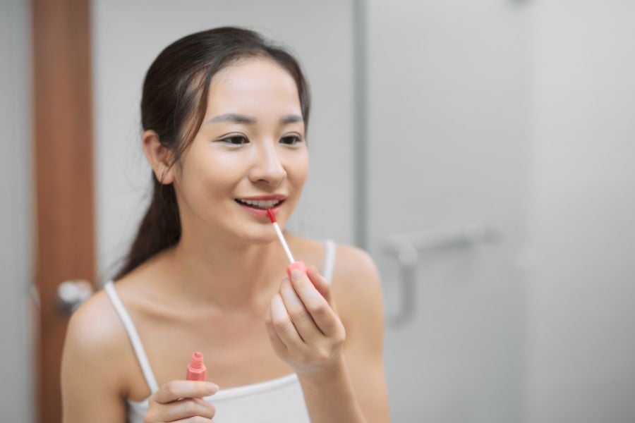 Người phụ nữ đang thoa son môi trước gương trong phòng tắm, mặc áo hai dây màu trắng.