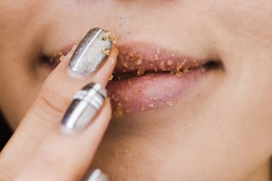 Người đang thoa hỗn hợp tẩy tế bào chết lên môi, sử dụng ngón tay với móng được sơn màu bạc.