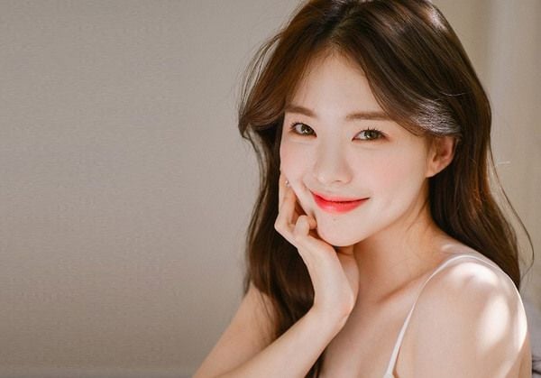 Người phụ nữ ngồi tựa cằm lên tay, mỉm cười nhìn về phía máy ảnh trong ánh sáng tự nhiên nhẹ nhàng.