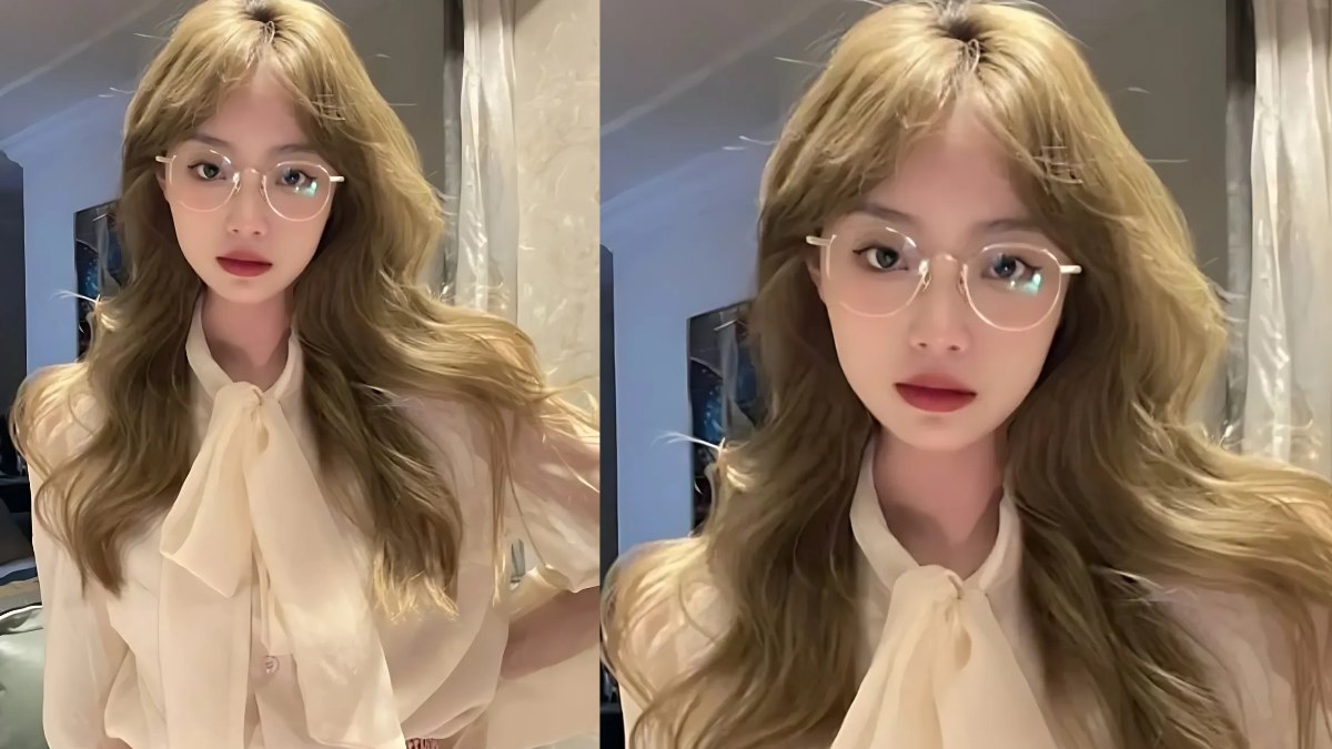 nau-kiwi-6 Một người đeo kính, tóc dài, mặc áo sơ mi sáng màu đứng trong phòng có rèm cửa.