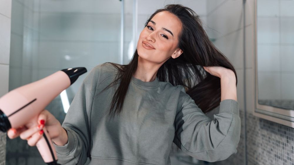 Người phụ nữ sấy tóc trong phòng tắm, mặc áo dài tay màu xám, cầm máy sấy màu hồng nhạt.