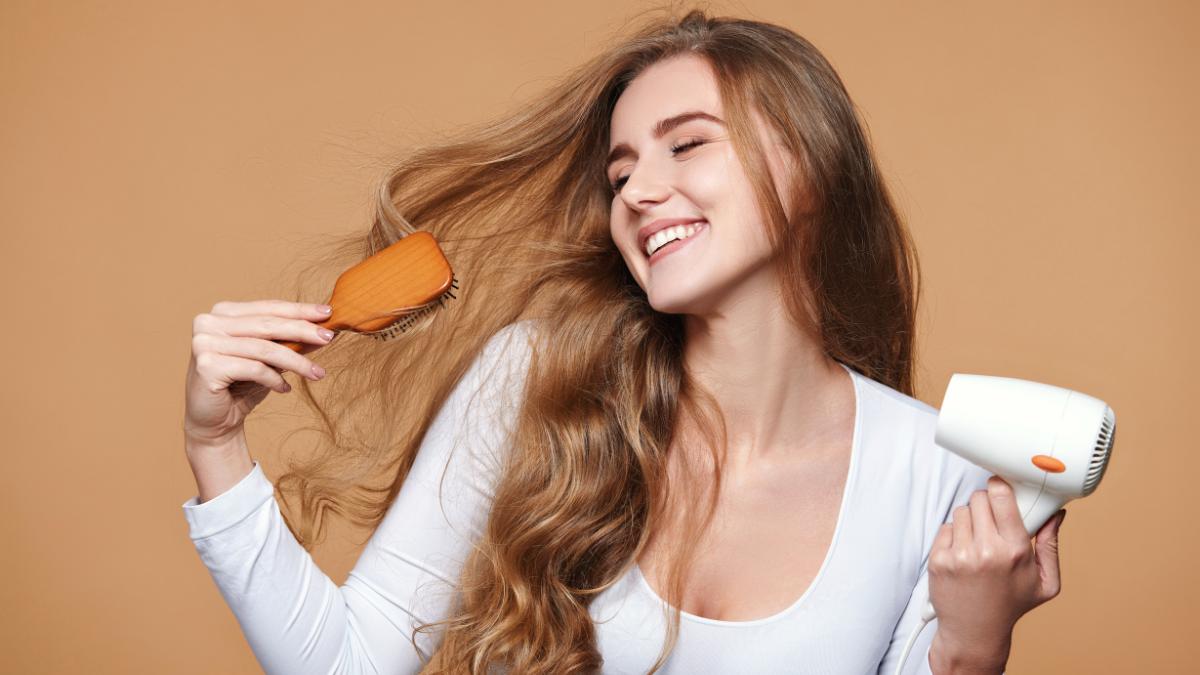 Người phụ nữ cầm máy sấy tóc và lược, mỉm cười thoải mái với mái tóc dài, nền màu nâu nhạt.