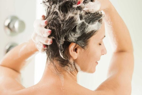 Người đang gội đầu với nhiều bọt xà phòng, tay xoa đều trên tóc, nền trắng sáng.