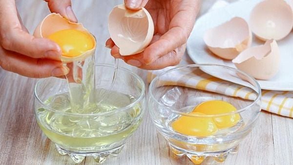 Hai tay tách lòng đỏ và lòng trắng trứng vào hai bát thủy tinh trên bàn gỗ, với vài vỏ trứng xung quanh.