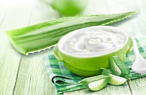 Bát sữa chua trên khăn kẻ caro xanh, kèm lá và miếng nha đam tươi trên nền gỗ nhạt màu.