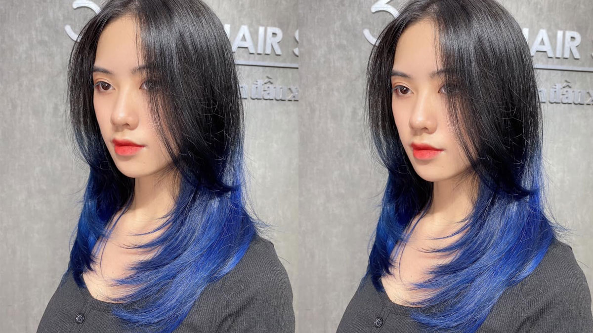 Người mẫu với tóc đen dài, phần đuôi nhuộm xanh, đứng trước nền tường xám.