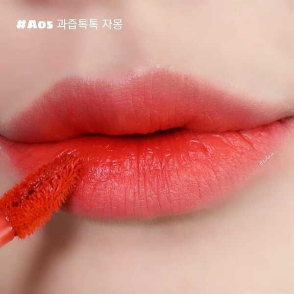 Đôi môi được tô son màu đỏ cam, cọ son đang chạm vào môi.