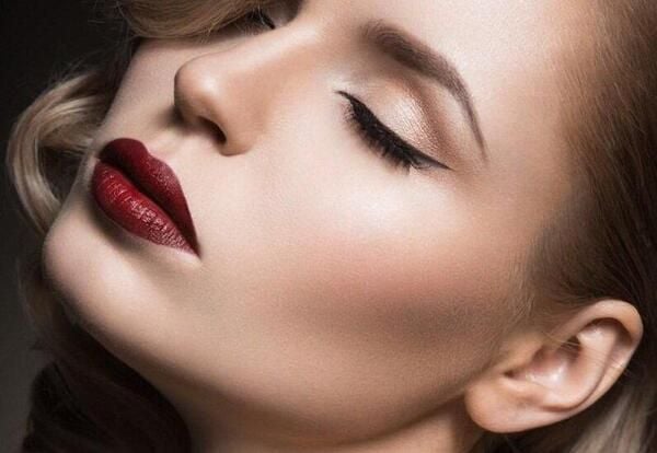 Gương mặt người với lớp trang điểm tông màu trung tính, môi đỏ đậm, mắt nhắm, tóc xoăn nhẹ.