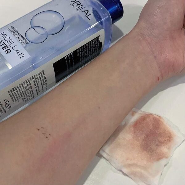 rang_dang_mong_doi_nho_cong_nghe_mixen_loai_bo_bui_ban__lop_trang_diem_6fd929143df242d2b74ed835fa58da27_grande Cánh tay với chai nước tẩy trang và bông tẩy trang có vết bẩn màu nâu đỏ trên nền trắng.