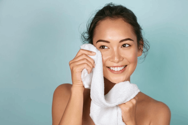 Người phụ nữ mỉm cười, dùng khăn trắng lau mặt, nền xanh nhạt.