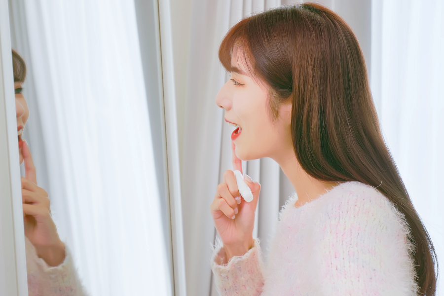 Người phụ nữ trẻ cầm son dưỡng, áp sát gương, mặc áo len trắng, đang cười trước gương trong phòng sáng.