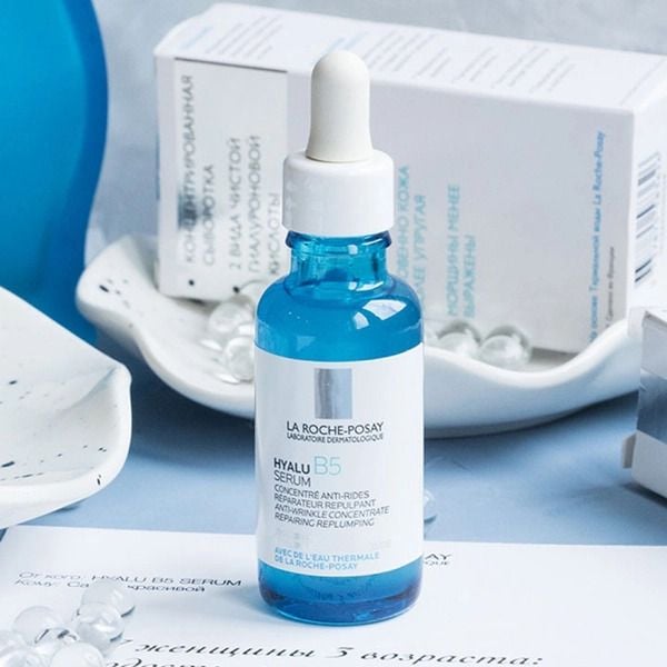 Chai serum màu xanh với nắp nhỏ giọt, đặt trên bàn cạnh hộp và đĩa trang trí.