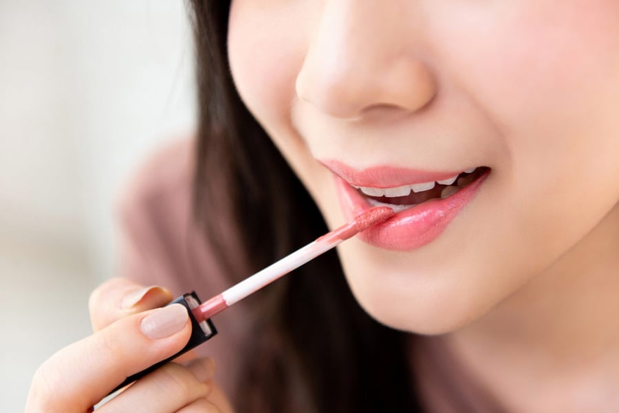 Người đang thoa son môi màu hồng nhạt bằng cọ, môi mỉm cười nhẹ, nền mờ.