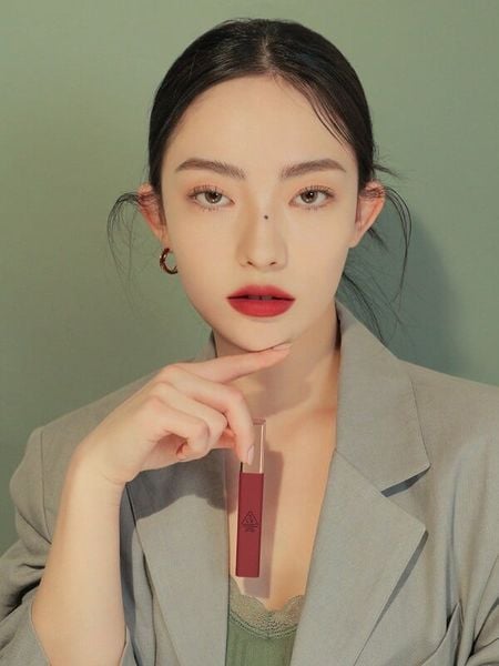 Người phụ nữ mặc áo vest cầm thỏi son đỏ, nền xanh lá.