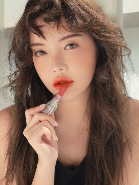 Người phụ nữ cầm thỏi son màu cam, thoa lên môi, tóc xoăn nhẹ, trang điểm tự nhiên, ánh sáng tự nhiên chiếu vào.