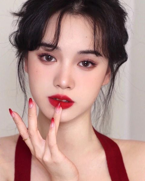 Người phụ nữ với tóc đen, trang điểm môi đỏ và mắt đậm, chạm nhẹ vào môi bằng ngón tay, nền trắng.
