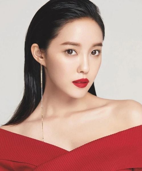 Người phụ nữ mặc áo đỏ, tóc đen dài, đeo khuyên tai, trang điểm nhẹ với son môi đỏ, nhìn thẳng vào máy ảnh.