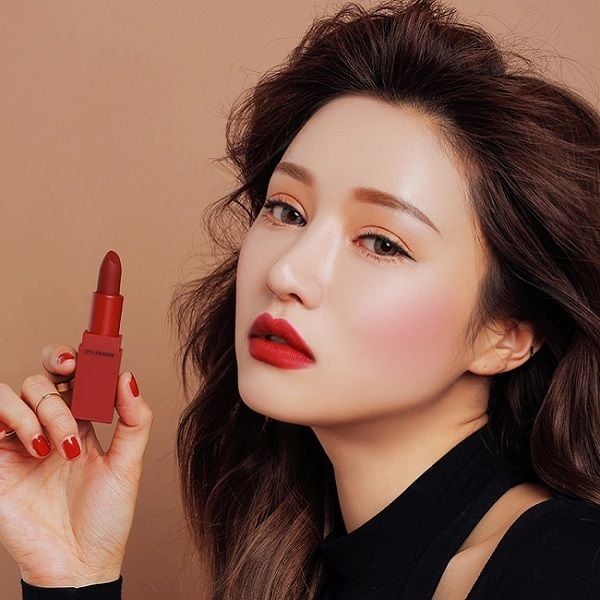Người phụ nữ cầm thỏi son đỏ, trang điểm tinh tế, tóc dài, trên nền màu nâu.