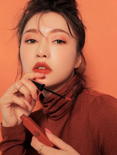 Người phụ nữ cầm thỏi son màu cam, tay cầm cọ son, nền màu cam, trang điểm tông cam hài hòa.
