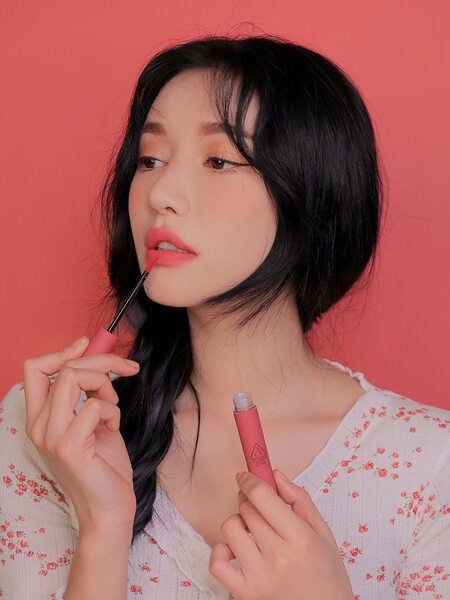 Người phụ nữ cầm cọ thoa son môi màu hồng, nền hồng, mặc áo trắng hoa nhí.