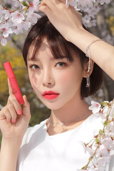 Người phụ nữ cầm son môi màu đỏ đứng giữa khung cảnh hoa anh đào nở rộ.