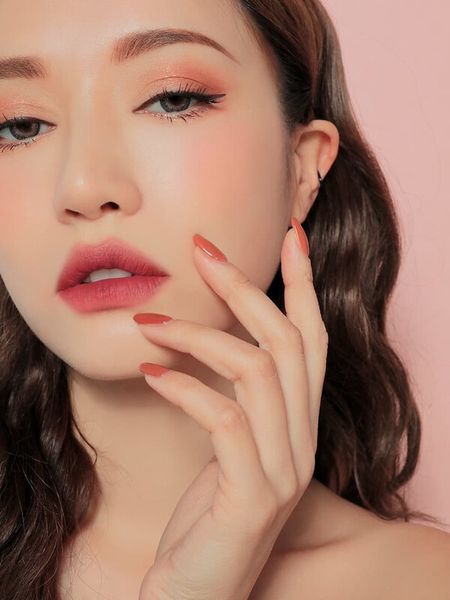 Người phụ nữ với trang điểm tự nhiên, tóc dài, móng tay sơn đỏ, nền màu hồng nhạt.