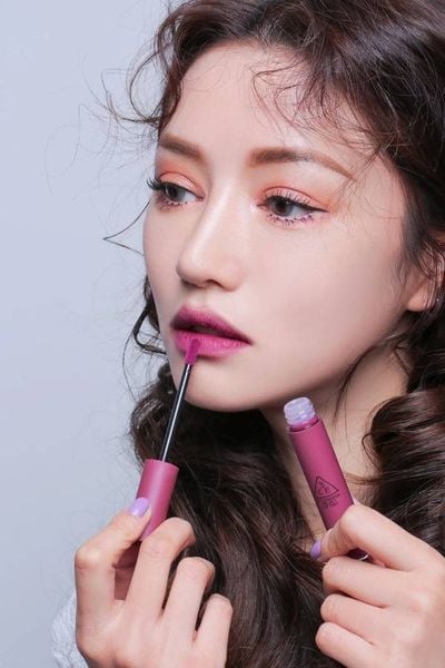 son_mau_hong_tim_la_su_ket_hop_giua_tone_hong_va_tim_2441a1d177144c01930a619550d34c97_grande Người phụ nữ đang tô son màu hồng tím lên môi, tay cầm cọ son, nền trắng.