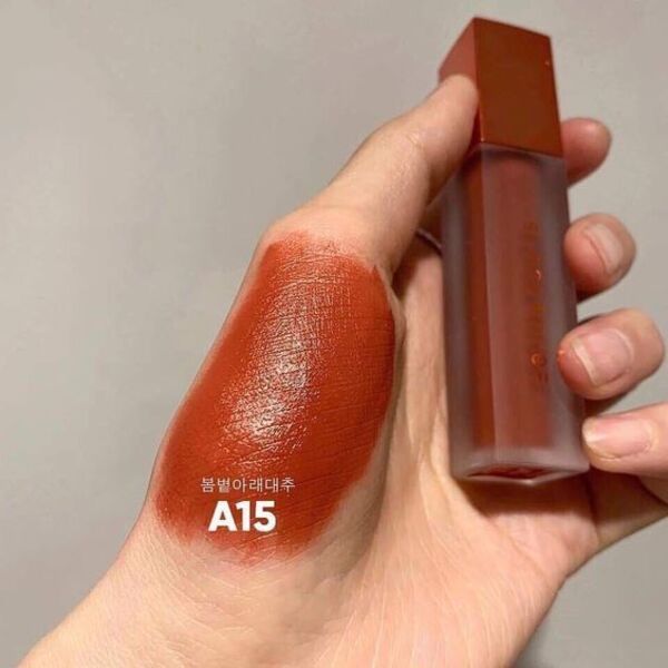 Một tay cầm lọ son màu nâu đỏ, với màu son được thử trên mu bàn tay bên cạnh mã A15.