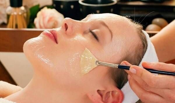 Người đang được đắp mặt nạ dưỡng da tại spa, với tay cầm cọ thoa nhẹ lên mặt, xung quanh có dụng cụ làm đẹp.
