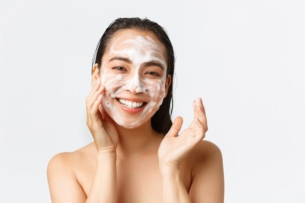 Người phụ nữ cười tươi, mặt có bọt sữa rửa mặt, nền trắng.