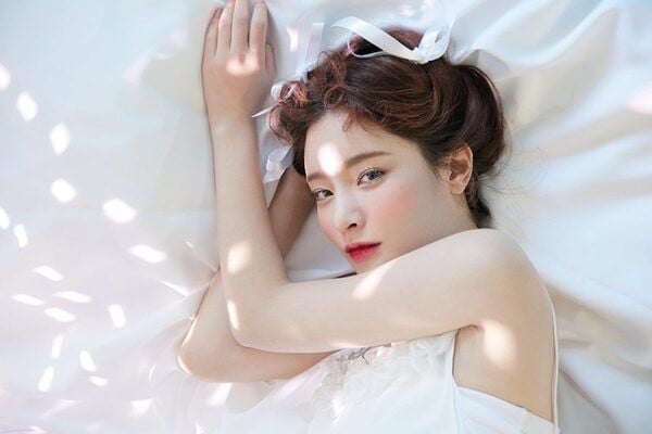 Người nằm trên giường với ánh sáng chiếu qua, mặc váy trắng, tóc búi và trang điểm nhẹ nhàng.