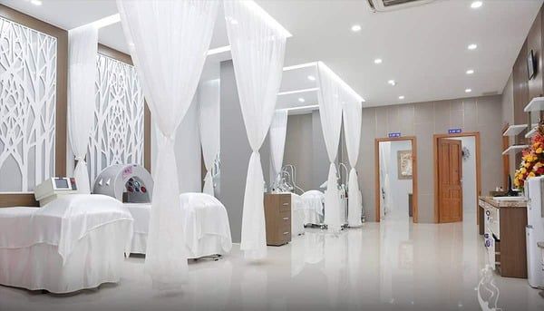 Phòng spa hiện đại với giường trị liệu, rèm trắng và thiết bị chăm sóc da, ánh sáng dịu nhẹ.